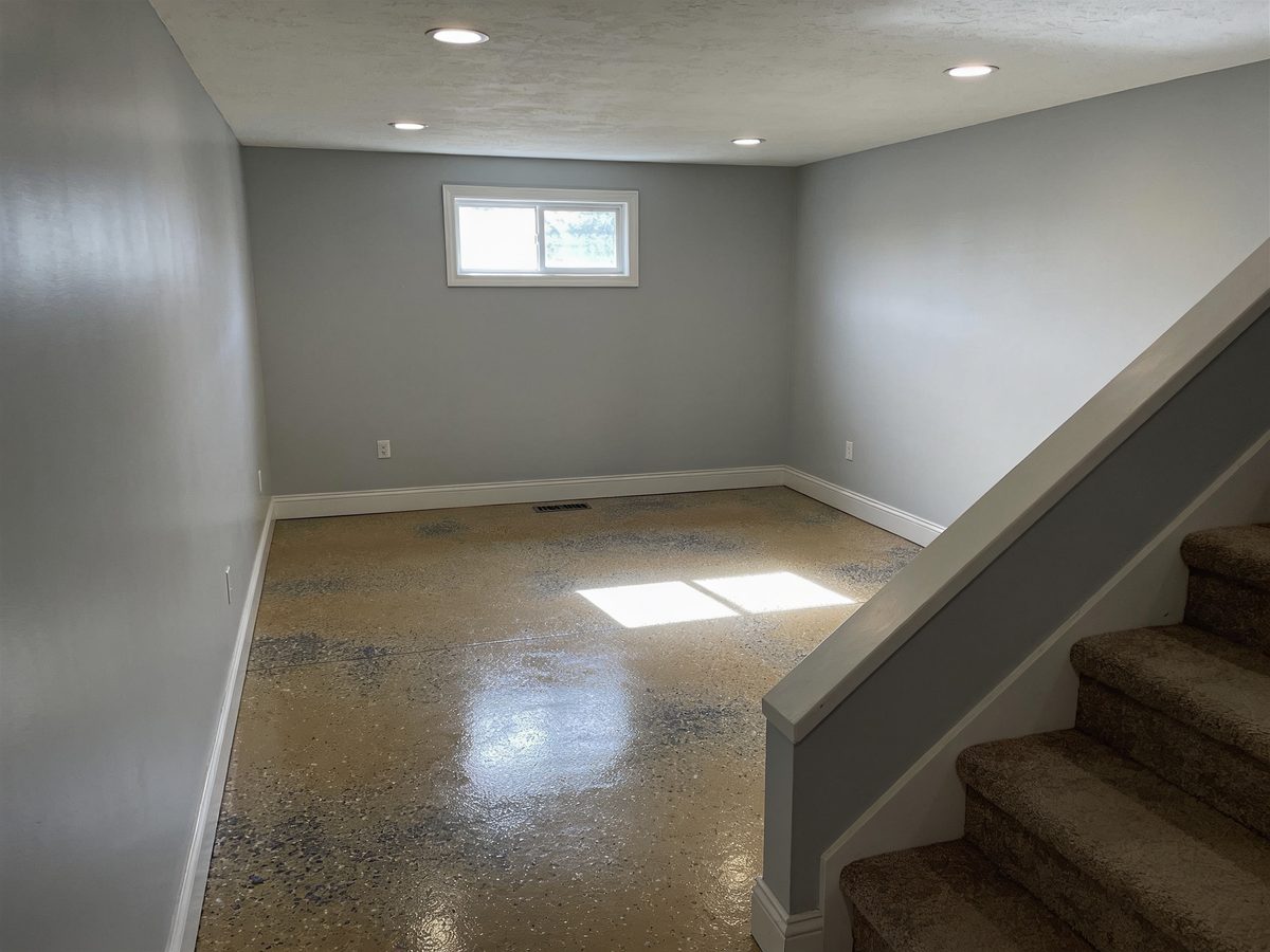 A finished basement room with a tan and charcoal flake floor coating and a stair descending into the room